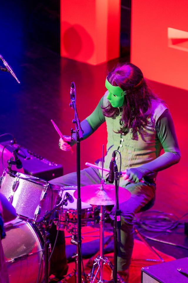 The/Theys drummer in a green mask playing the kit under red stage light