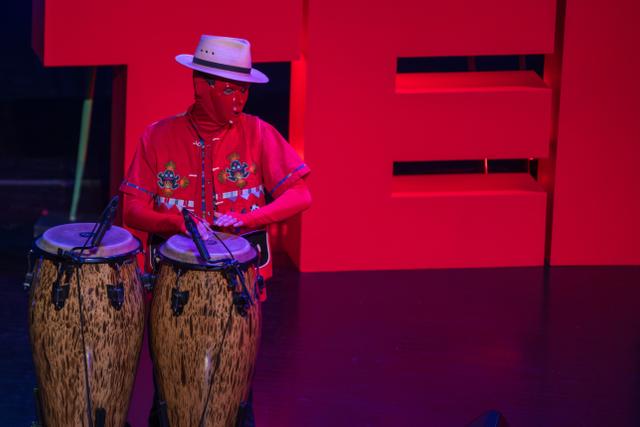 The/Theys percussionist in a red mask and white hat playing congas in front of the TEDx letters
